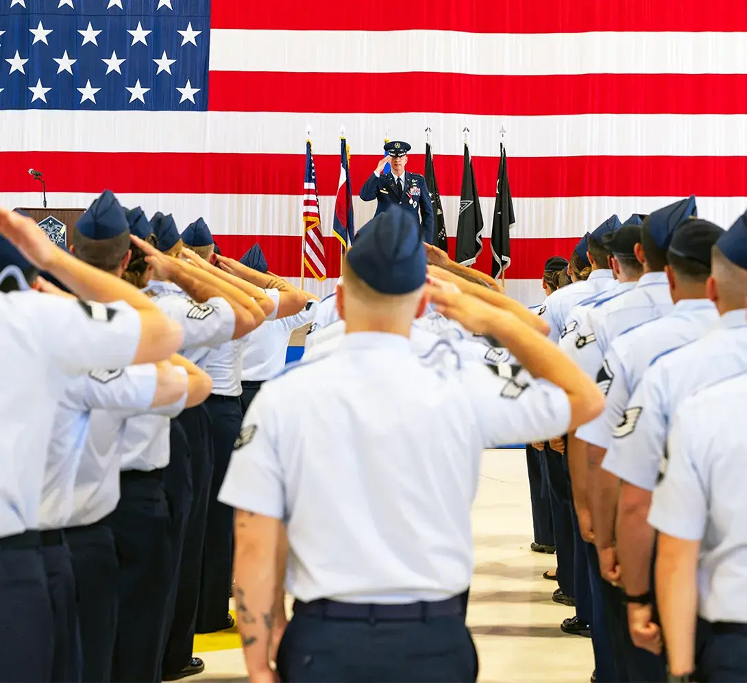 A group of U.S. service members in uniform salute an officer standing on stage in front of a large American flag and several military banners.